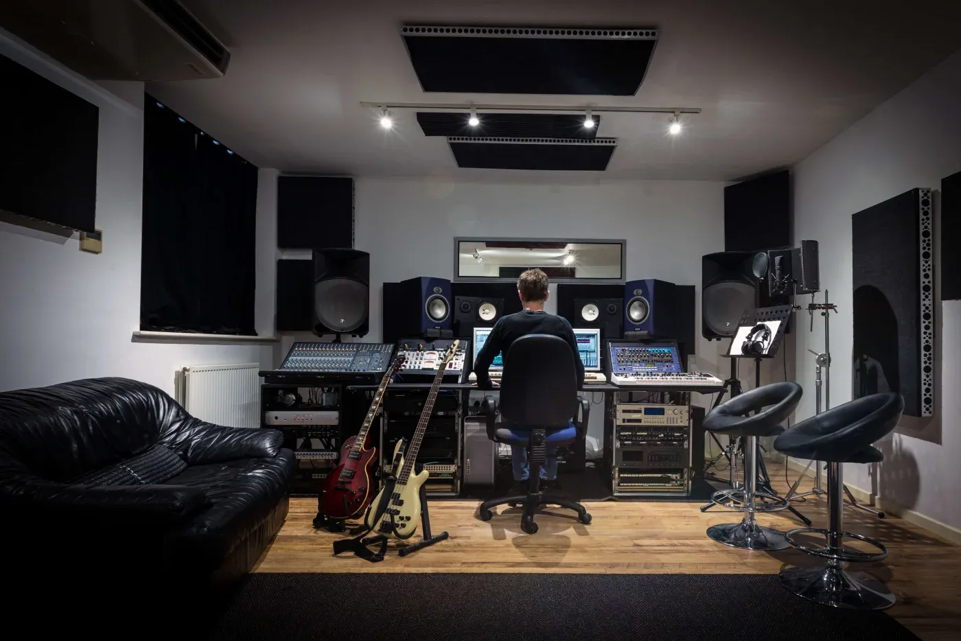 A person sits at a mixing desk in a recording studio with large speakers and monitors. Guitars rest against the desk, creating a focused, creative atmosphere.
