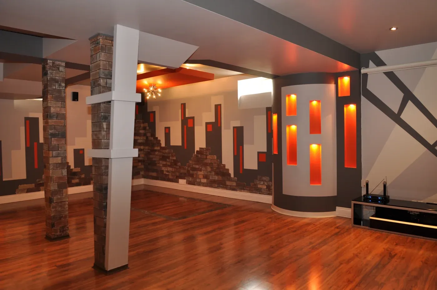 Modern room with wood floors, brick pillars, and geometric wall art in gray, orange, and white. Warm lighting creates a contemporary atmosphere.