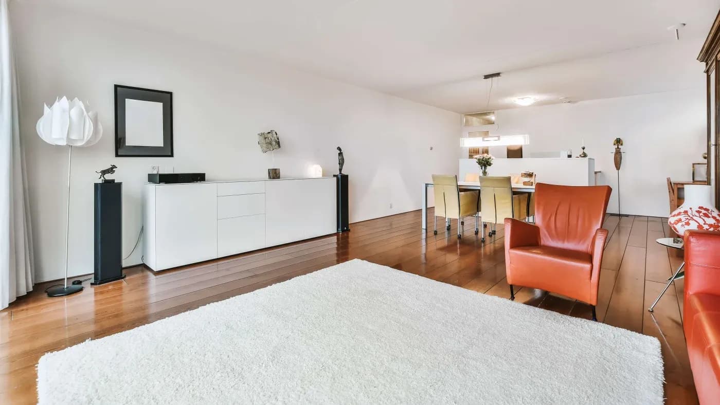 Spacious modern living room with wooden floors, a white rug, and red armchairs. Dining area with beige chairs; minimalist decor with contemporary art.