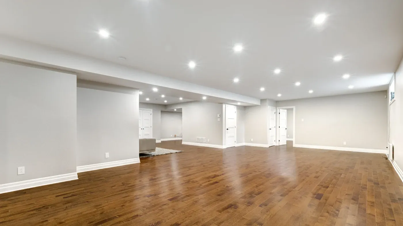 Spacious basement with polished wood floors, white walls, and recessed lighting. Minimalist design, empty except for a small gray rug.