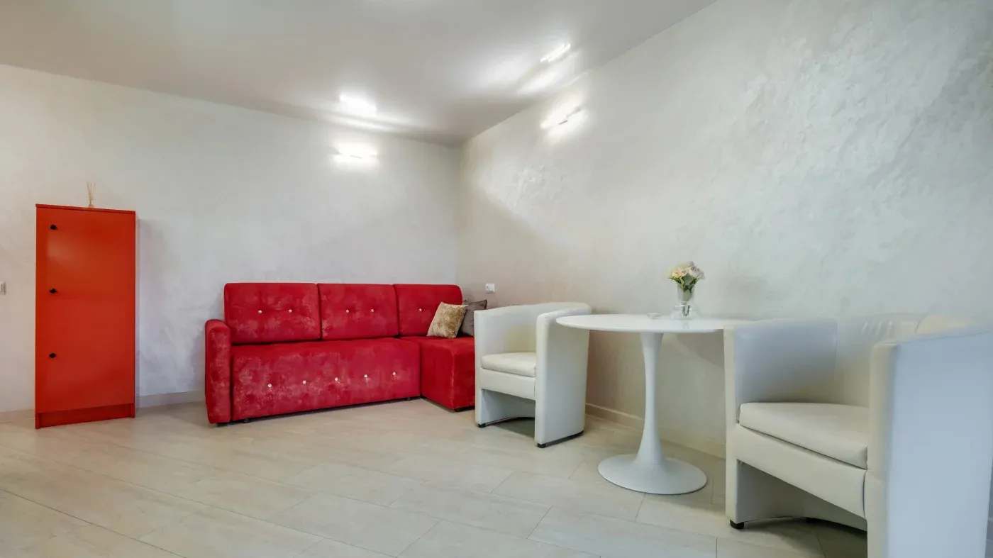 Modern room with a red couch and a matching cabinet on the left. Two white armchairs and a round table with a flower vase on the right. Neutral tones.