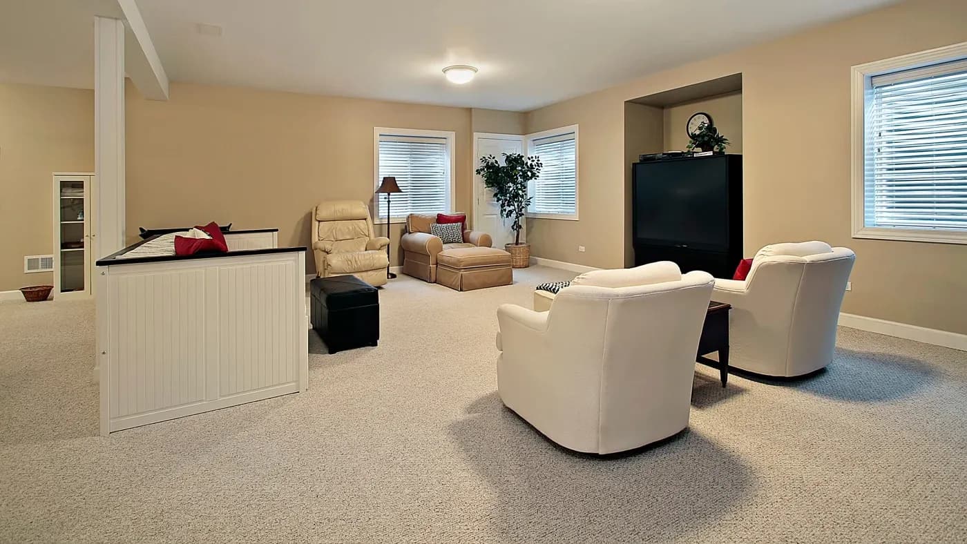 A spacious and cozy living room with beige carpet and walls. Two white armchairs face a large TV, while a tan sofa and recliner sit near a corner window.