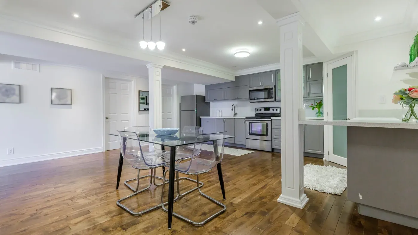 Modern kitchen and dining area with hardwood floors. Glass dining table with clear chairs, stainless steel appliances, and sleek gray cabinets. Bright, open space.