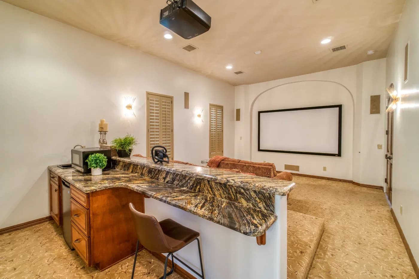 Home theater with a large screen framed by archway, plush brown sofas, and a marble bar with a stool, creating a cozy, luxurious atmosphere.