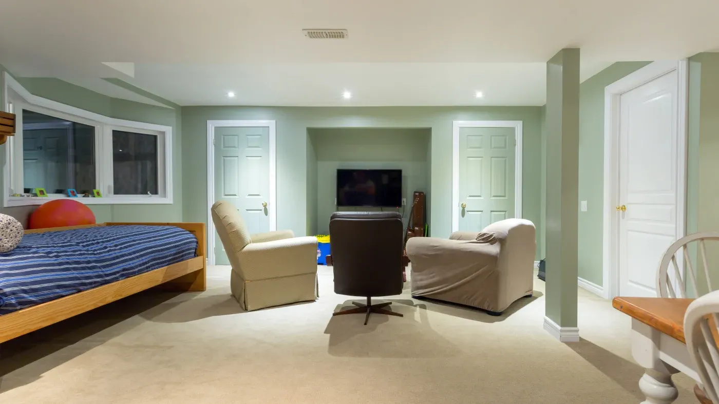 Cozy basement room with a bed, armchairs, and a TV. The walls are painted light green, creating a calm atmosphere. Bright, recessed lighting.