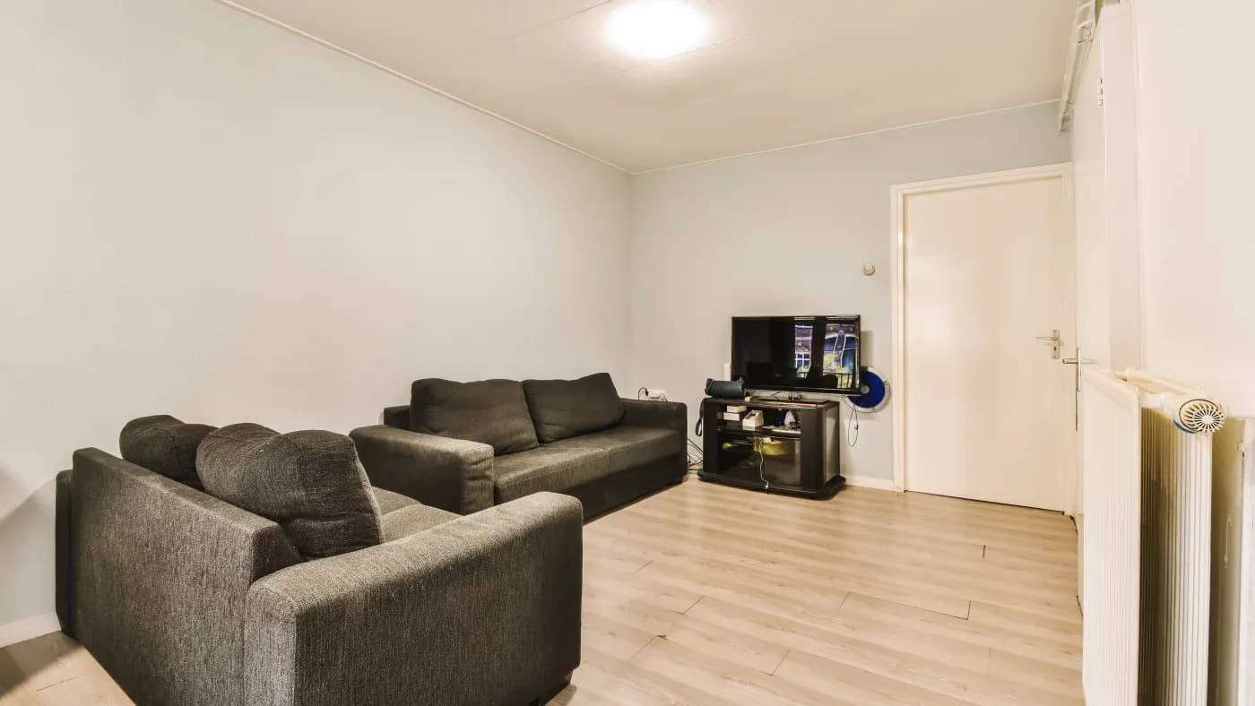 A minimalist living room with light wood flooring, gray walls, and two gray couches. A small TV sits on a black stand. The space feels simple and cozy.