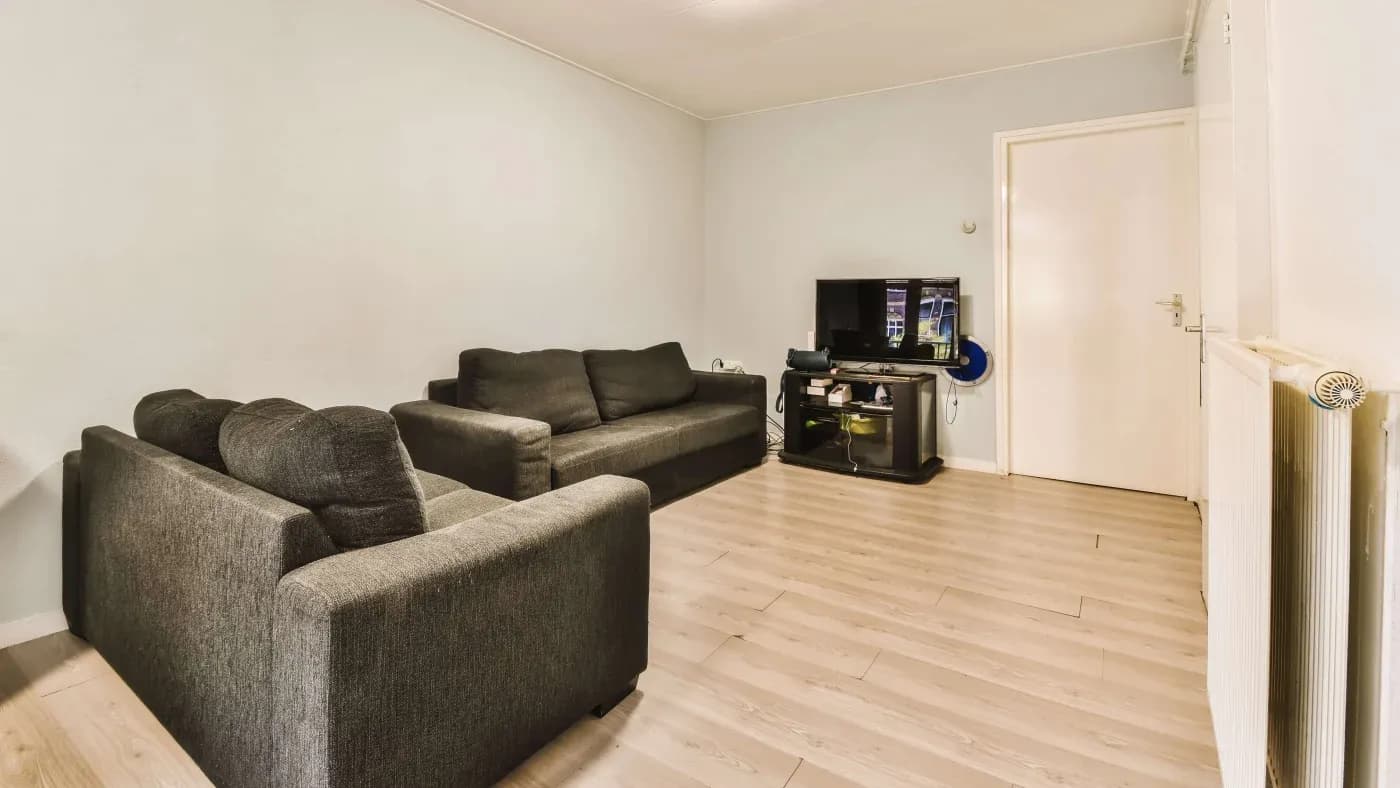 A minimalist living room with two dark gray sofas facing a TV on a black stand. The room features light wood flooring and pale walls, creating a calm atmosphere.