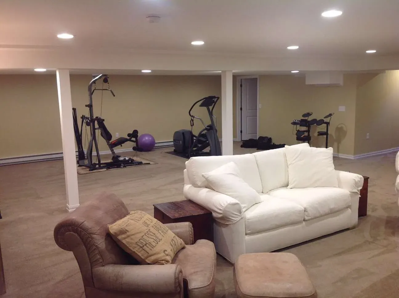 Basement room with white sofa, brown armchair, and ottoman on carpeted floor. Exercise equipment and a purple yoga ball are in the background.
