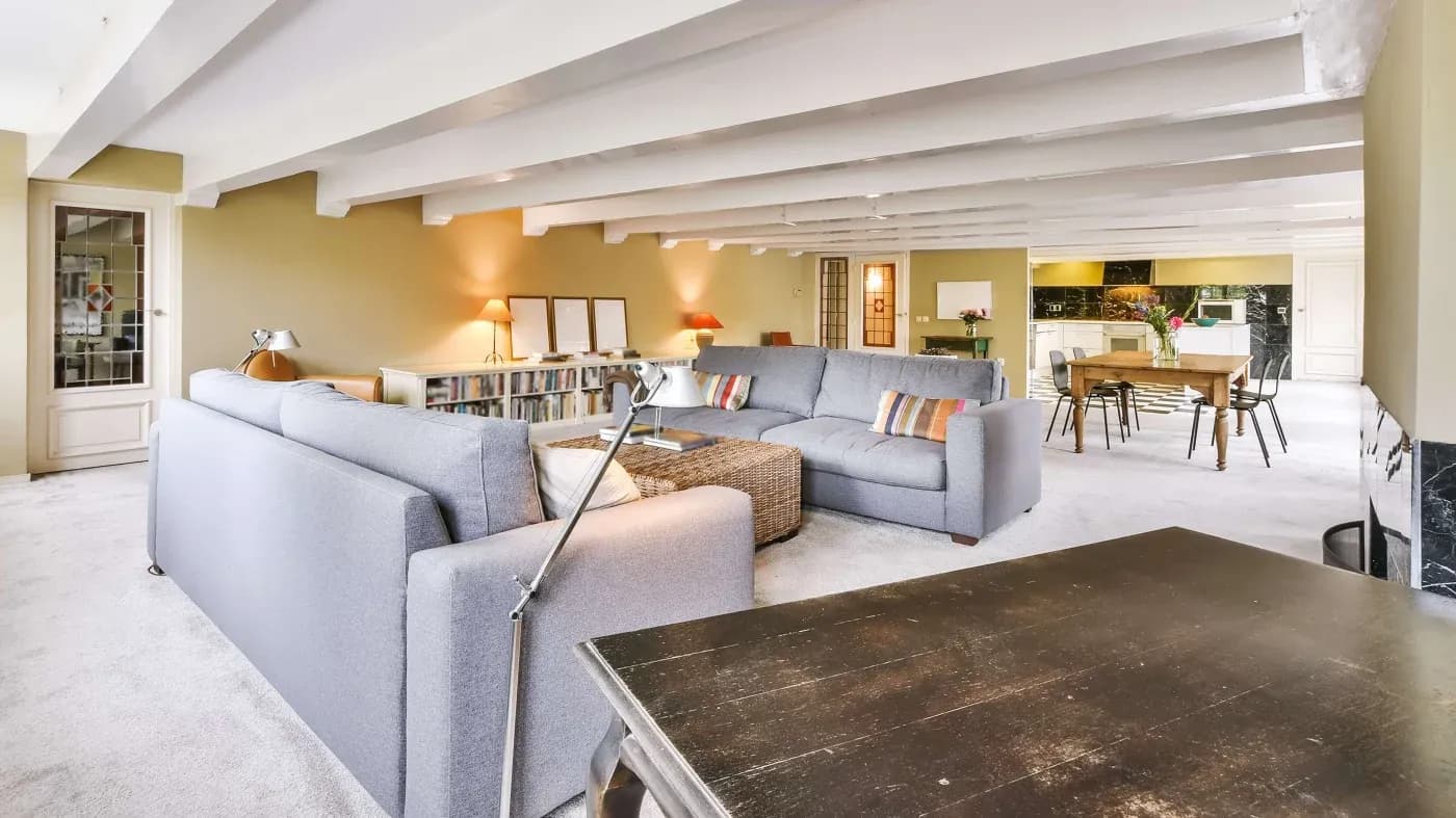 Spacious living room with light gray sofas, a wicker coffee table, and colorful cushions. Dining table and kitchen in the background, creating a cozy, inviting atmosphere.