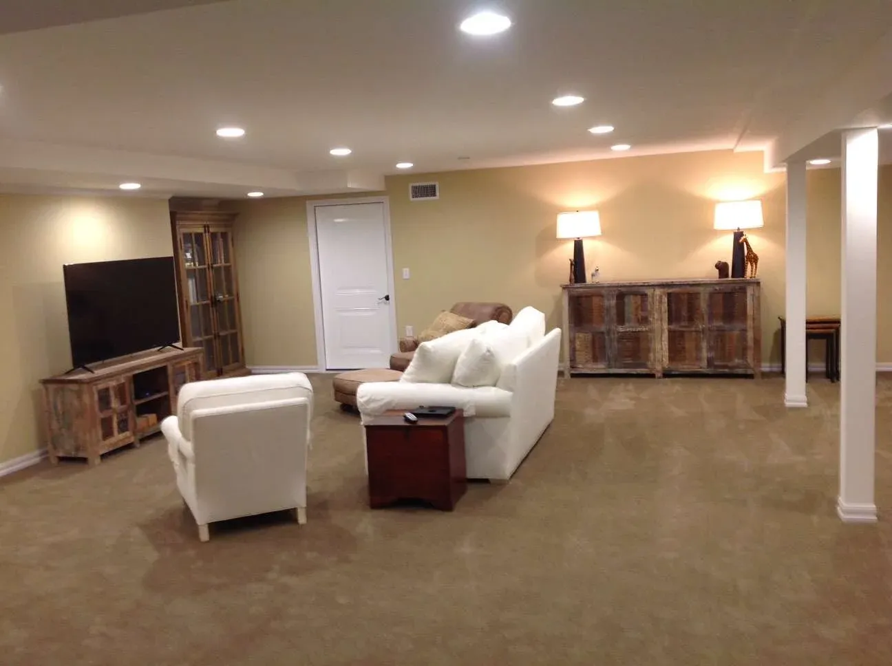 Basement room with beige walls and carpet, featuring white sofas and a large flat-screen TV. A wooden cabinet with lamps adds warmth and coziness.