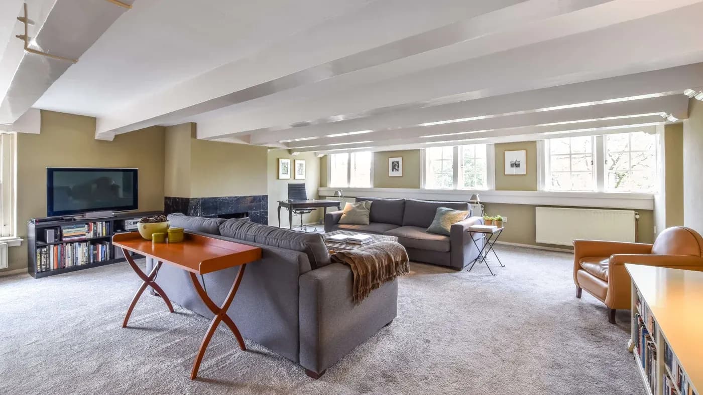 Spacious living room with gray sofas, a wooden table, a TV, and large windows letting in natural light. Cozy, modern decor with soft carpet.