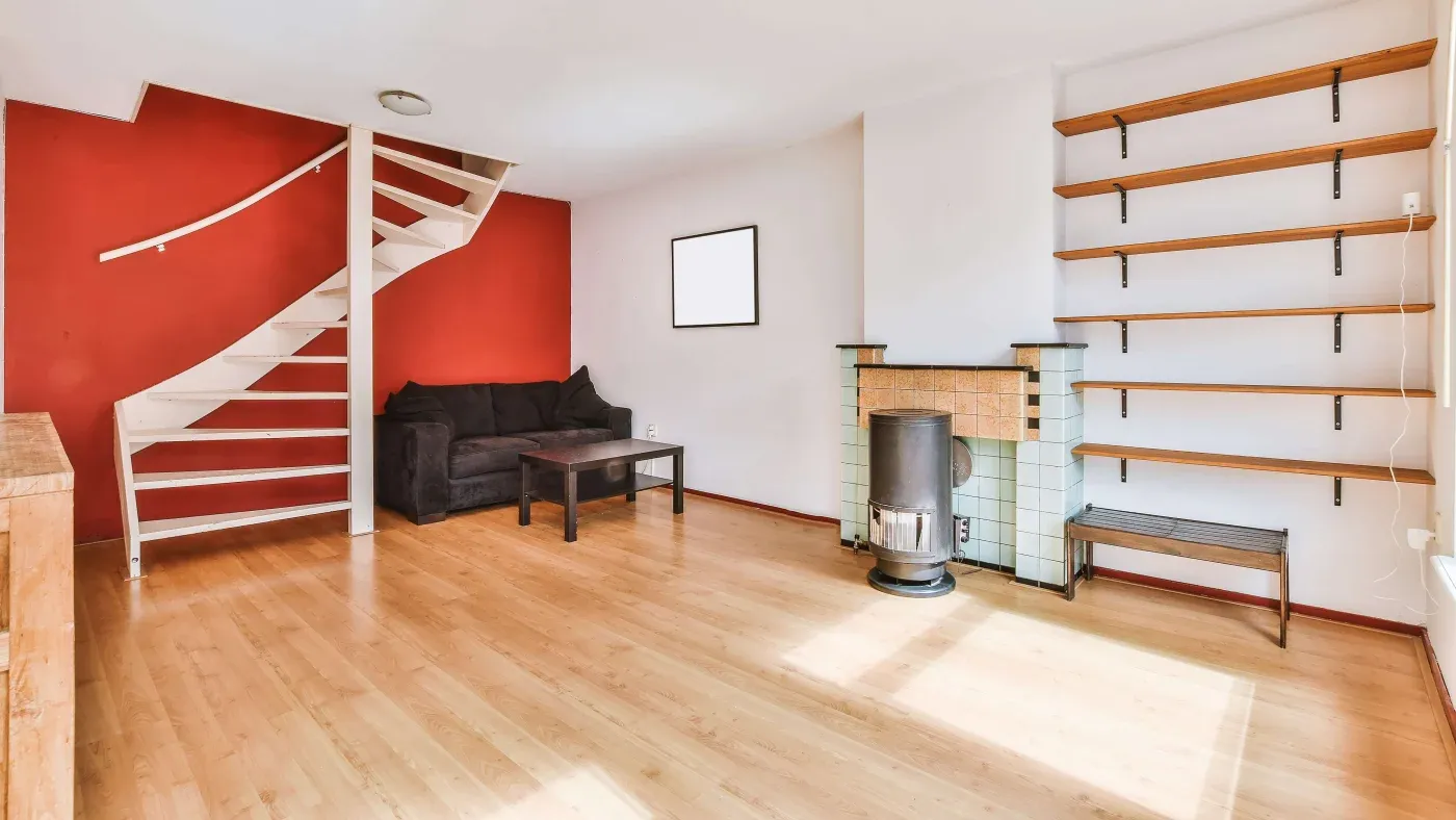 A cozy living room with red accent wall features a white spiral staircase, black sofa, wooden shelves, and a small wood-burning stove on light wood flooring.