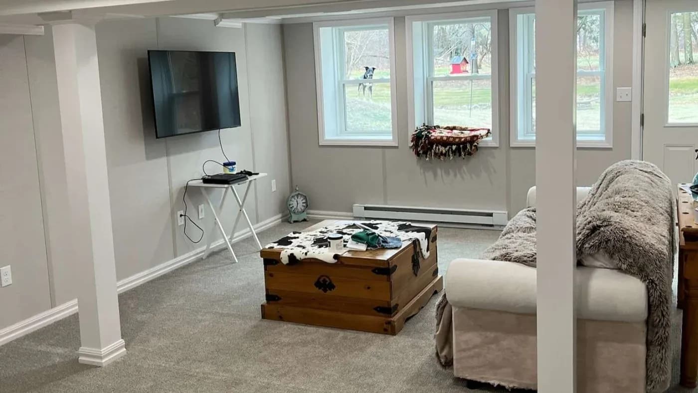 Cozy living room with a TV mounted on the wall, beige couch covered in a soft blanket, and a wooden chest as a coffee table. Large windows bring in natural light, revealing a view of the green lawn outside. The ambiance is warm and inviting.