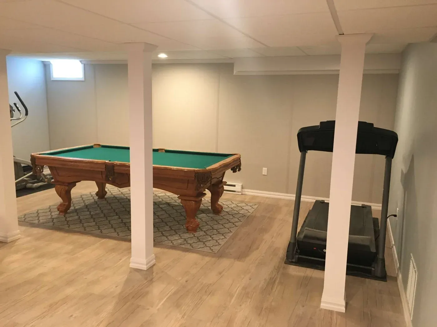 A cozy basement room features a pool table with a green felt top on a patterned rug and a treadmill nearby. Light wood flooring adds warmth.