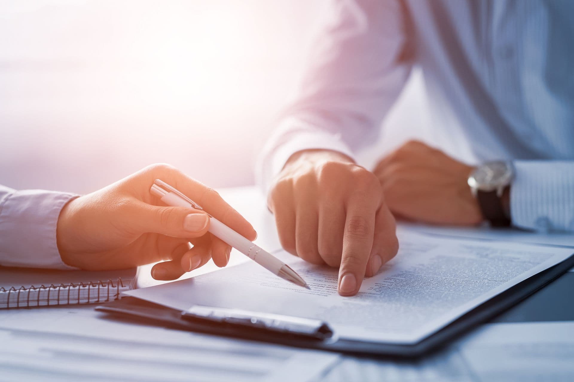 Two people in professional attire examine a document, one pointing while the other holds a pen. The setting is bright, suggesting focus and collaboration.