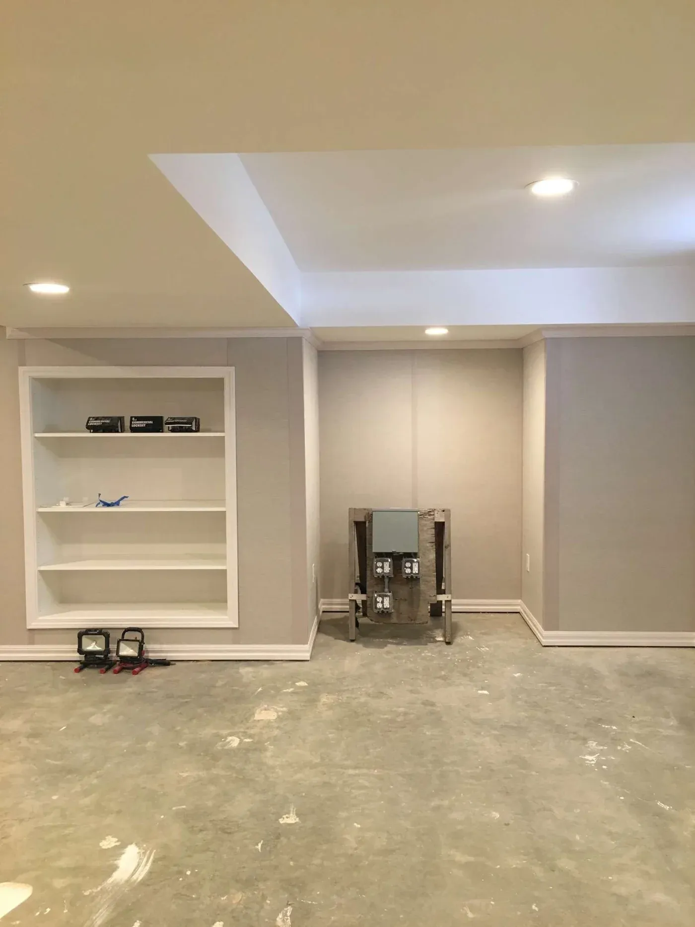 A partially renovated room with unfinished concrete floors, recessed lighting, and gray walls. Built-in white shelves hold small objects to the left.