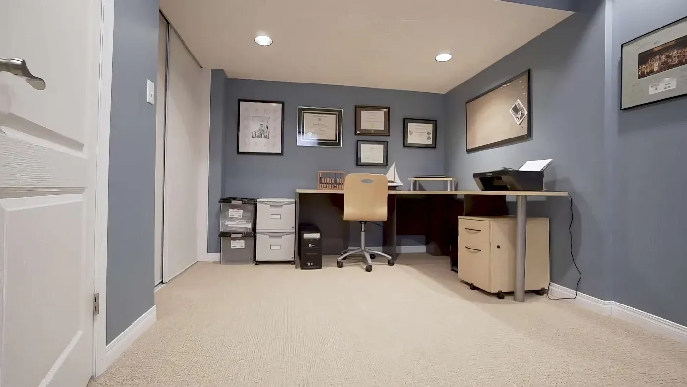 A home office with blue walls features a desk, beige chair, file cabinets, and a printer. Framed certificates and artwork adorn the walls, exuding a tidy, professional atmosphere.
