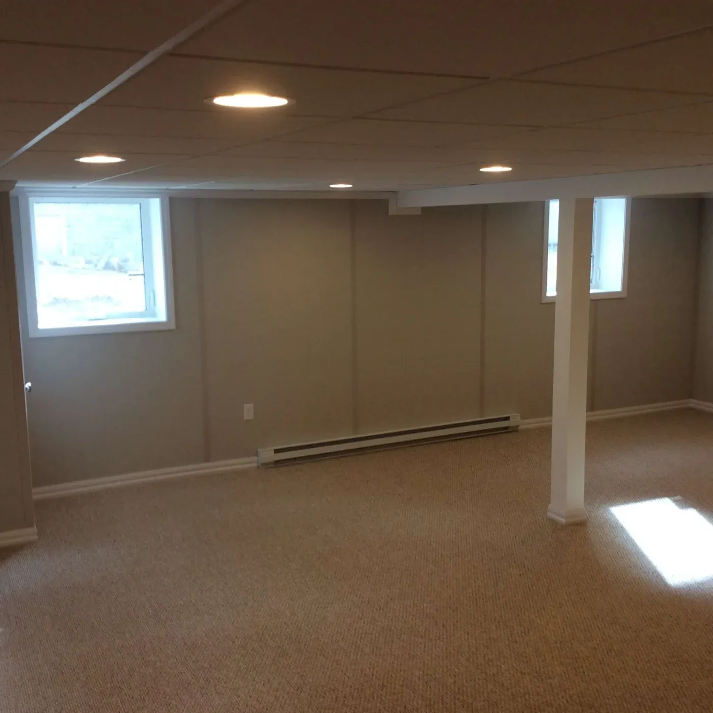 Empty basement room with beige walls, carpeted floor, support column, baseboard heater, recessed ceiling lights, and windows providing natural light