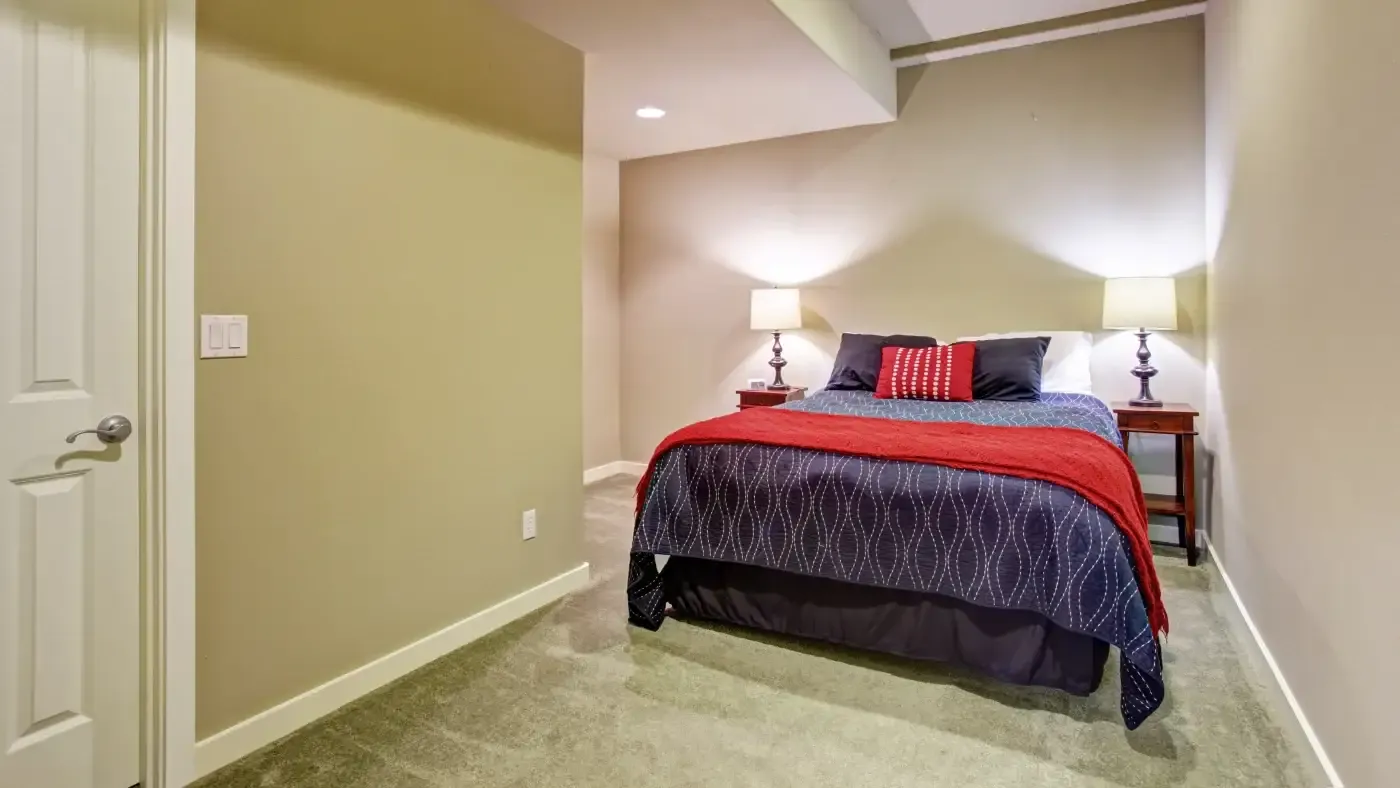 A cozy bedroom with beige walls, featuring a queen-sized bed covered in a red and navy blue patterned comforter. Two lamps on wooden nightstands emit soft light.