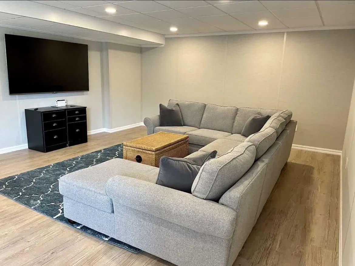 Spacious basement room with gray sectional sofa, wicker coffee table, large TV on the wall, and dark cabinet. Light wood floors and soft lighting create a cozy ambiance.