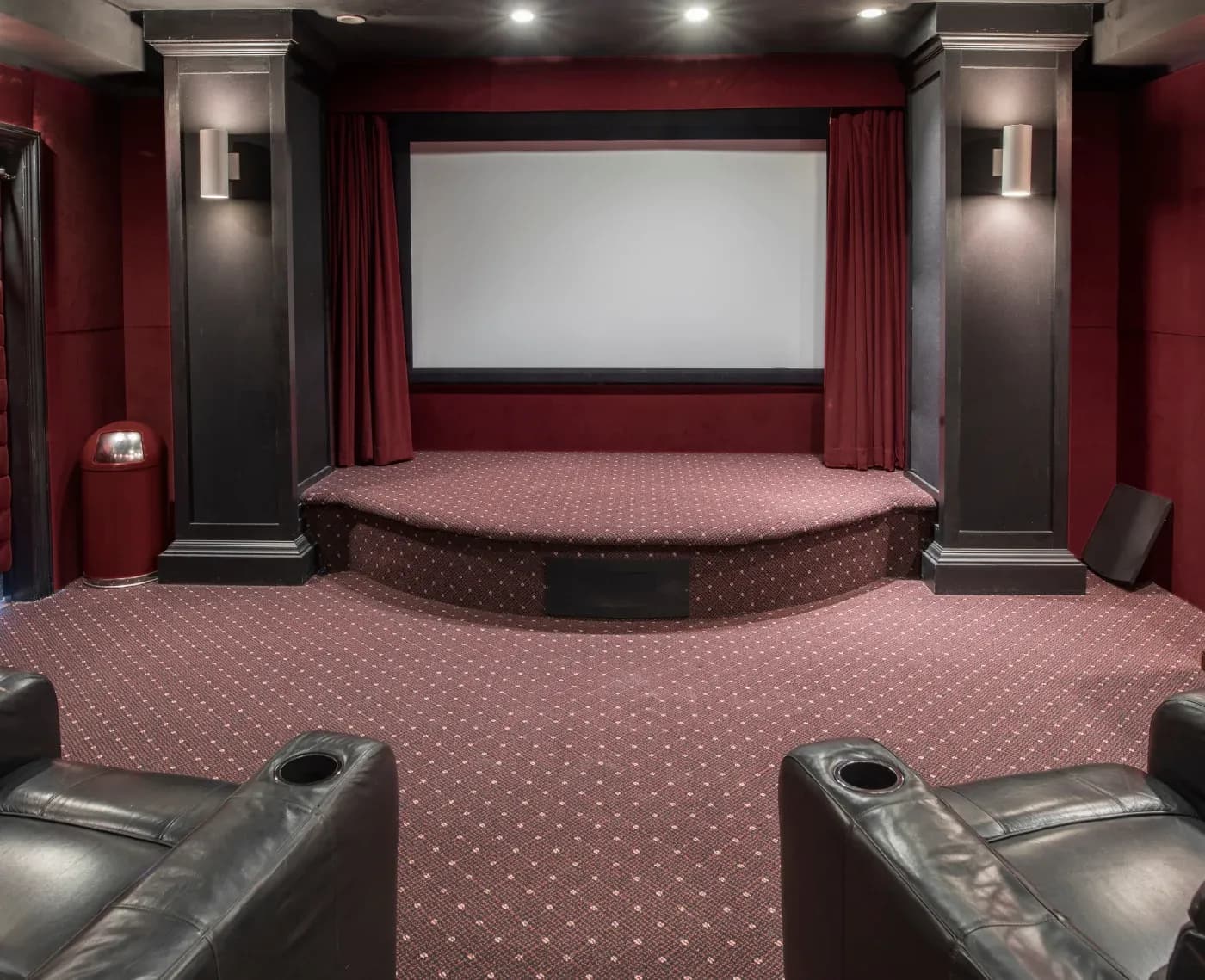 Elegant home theater with a large screen, red curtains, and plush burgundy carpet. Leather recliners offer comfort, creating a cozy atmosphere.