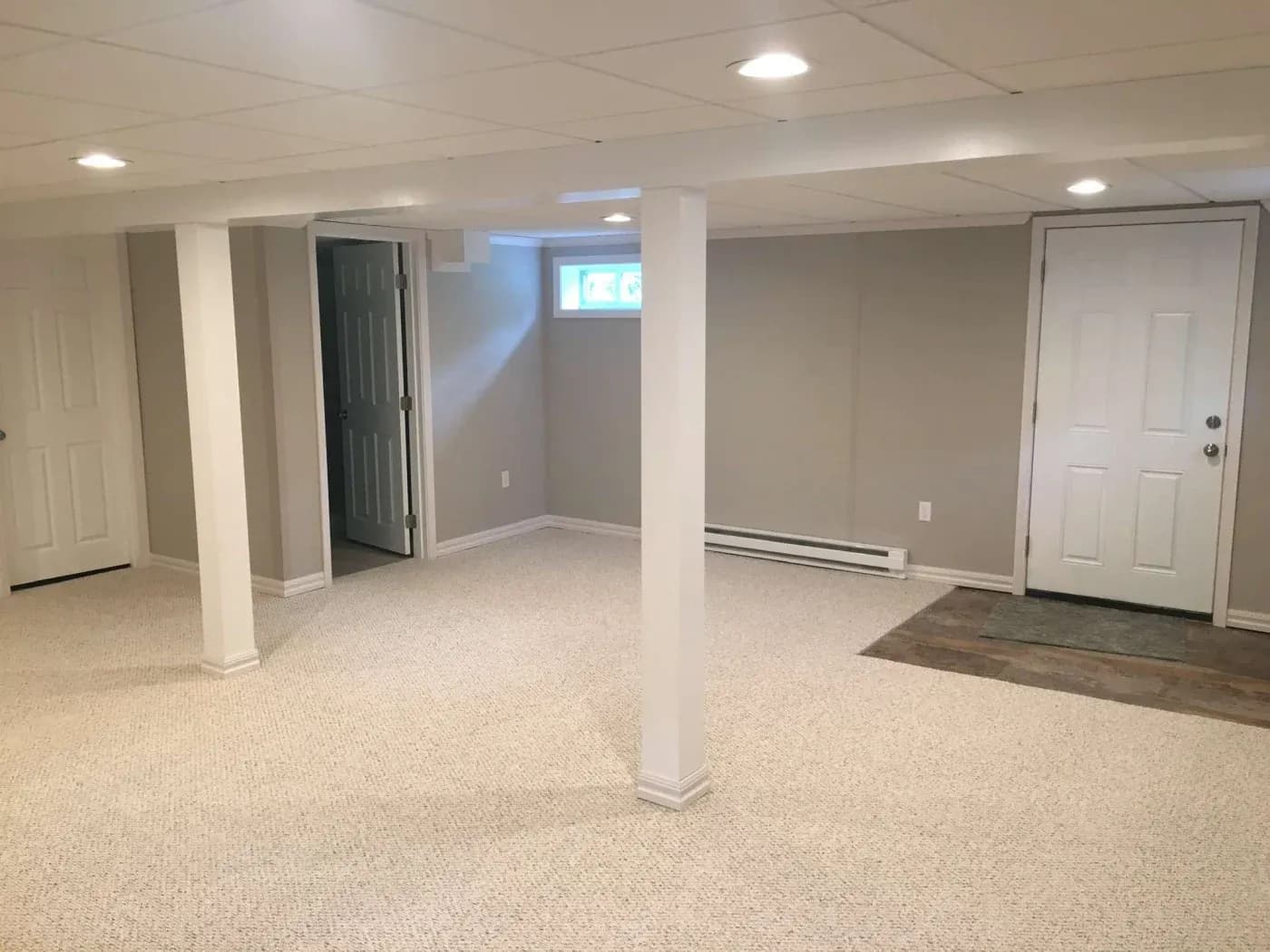 A finished basement with beige carpet, white walls, and ceiling beams. Two doors, a small window, and warm lighting create a spacious, welcoming feel.