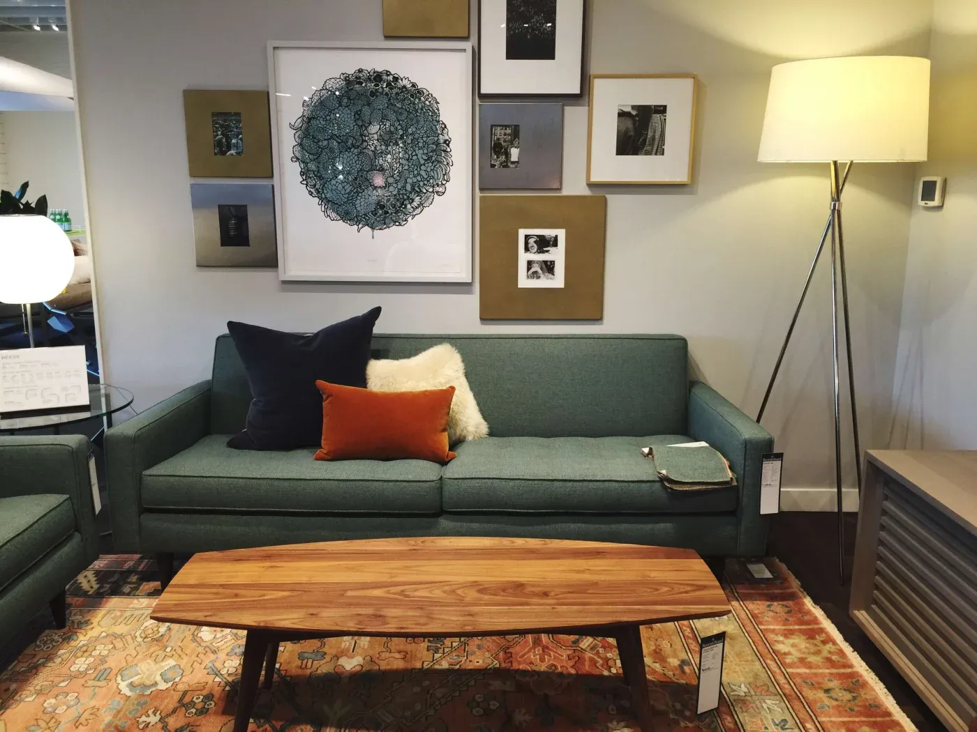 A cozy living room features a teal sofa with navy, orange, and white pillows. A wooden coffee table sits on a patterned rug. Art and a tall lamp give warmth.