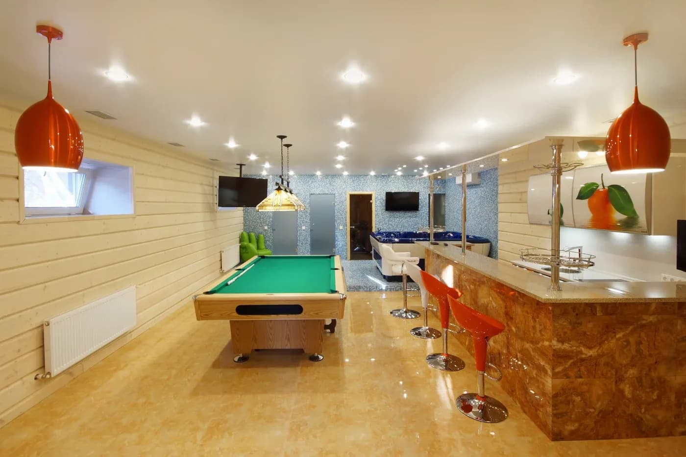Spacious modern rec room with pool table, glossy tiled floor, and orange pendant lights. Bar area with red stools and a large mural of oranges.