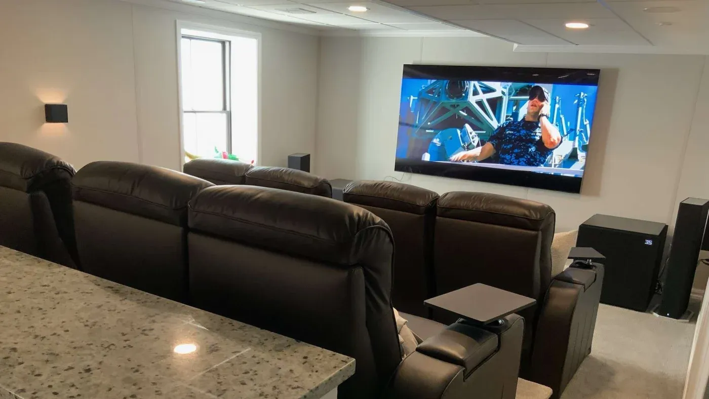 Home theater room with plush black leather recliners facing a large flat-screen TV showing a movie scene. Soft lighting and modern decor create a cozy atmosphere.