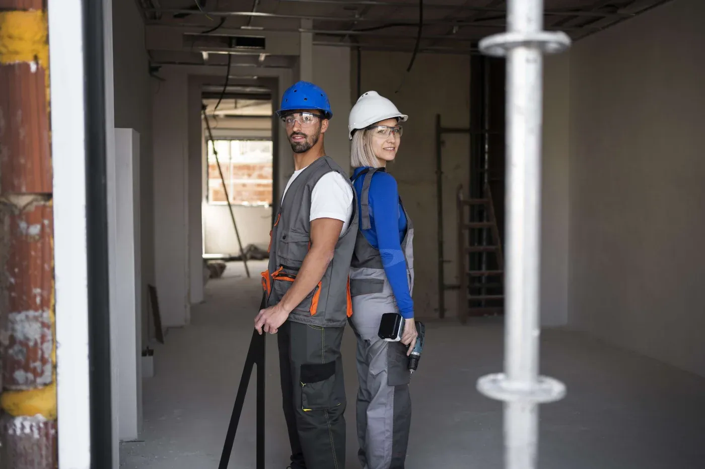 Two construction workers, a man and a woman, wearing hard hats and safety gear, stand back-to-back in a partially built room. They appear confident.