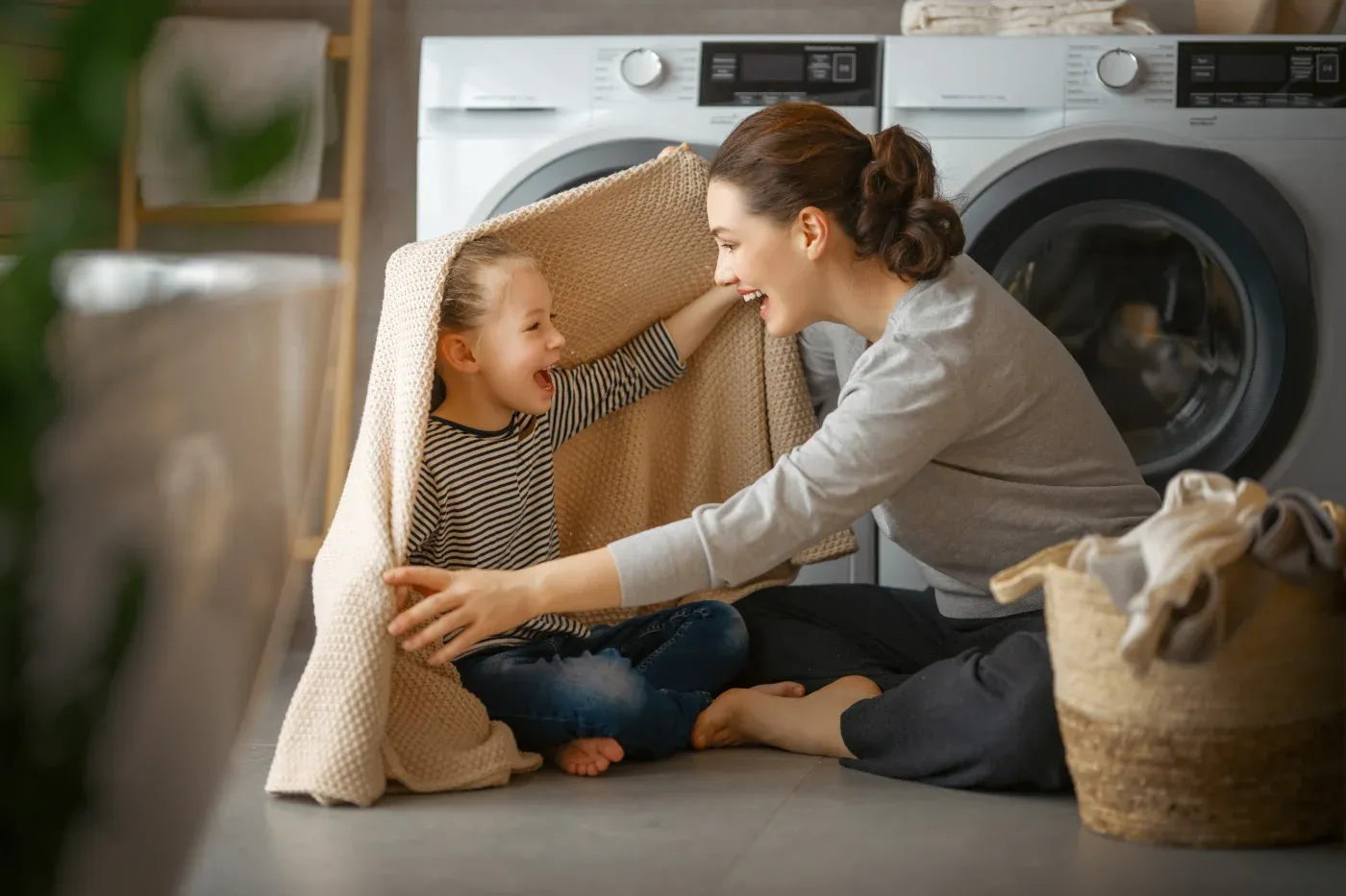 A mother and daughter playfully hide under a blanket in a laundry room. The setting is warm and joyful, with a basket of clothes nearby and washing machines in the background.