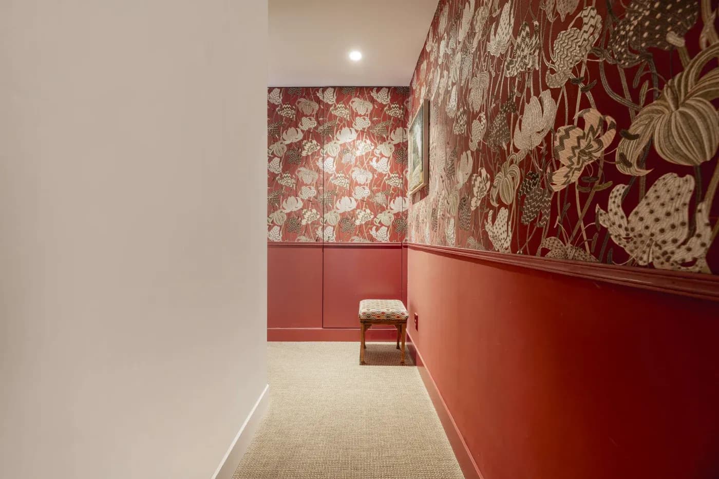 Narrow hallway with rich red walls and floral wallpaper, featuring an elegant upholstered bench under a spotlight, creating a cozy, vintage ambiance.