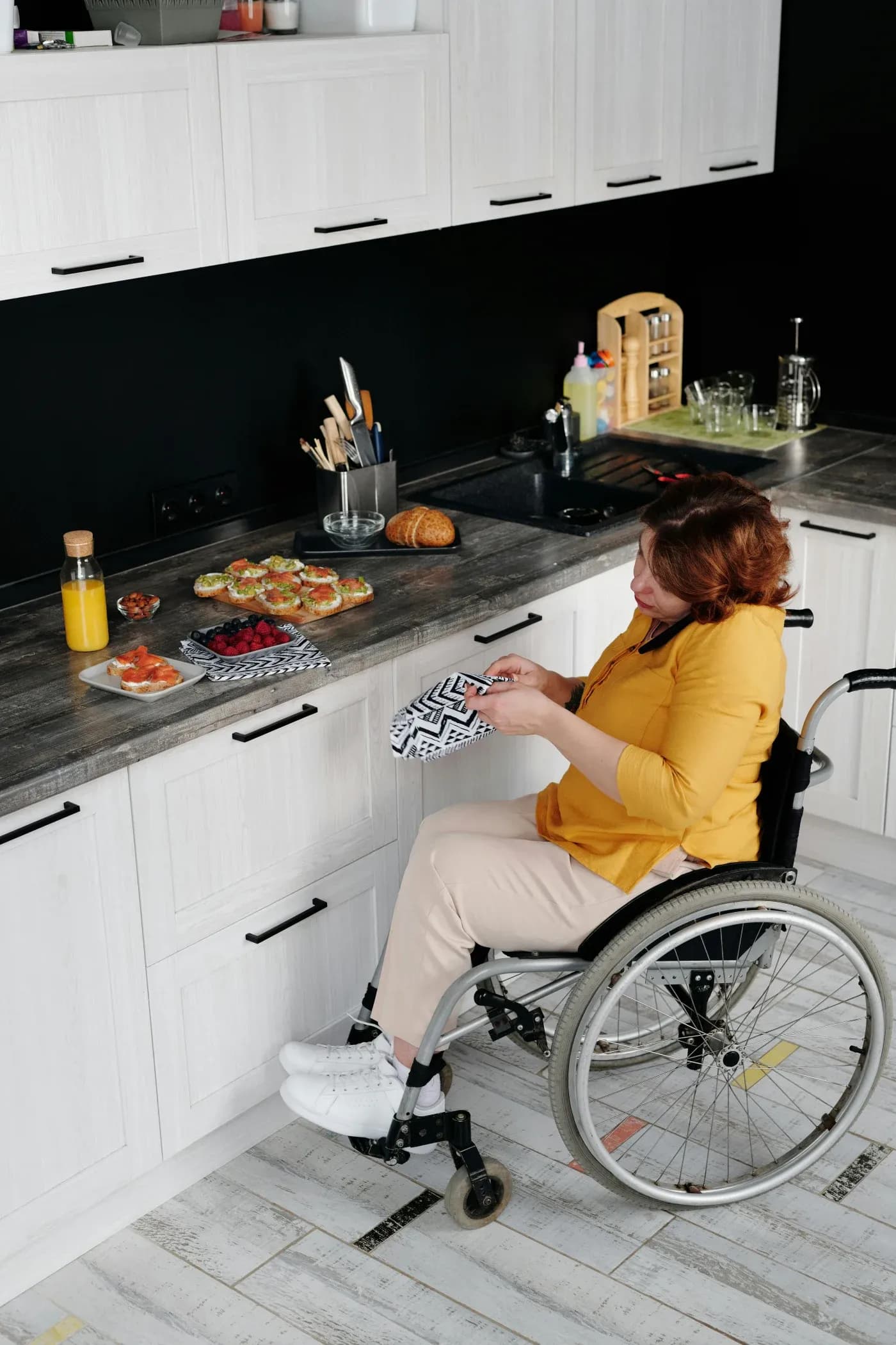 Woman in a wheelchair wearing a yellow shirt prepares food in a modern kitchen. The countertop has pastries, fruit, and juice, conveying a cozy atmosphere.