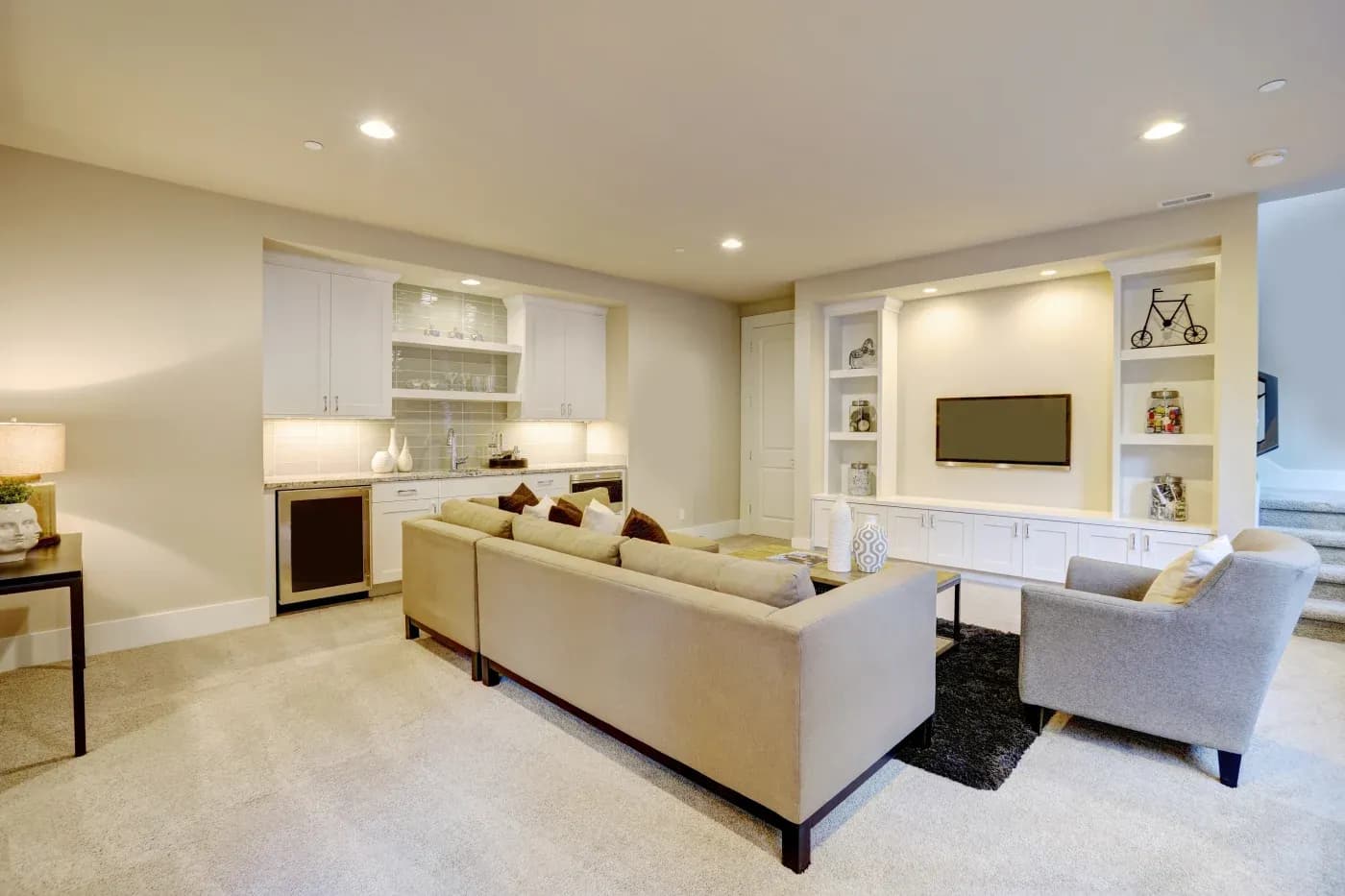 Modern living room with beige sofas and a gray armchair facing a wall-mounted TV. Built-in shelves surround the TV. A small kitchenette is visible. Relaxed ambiance.