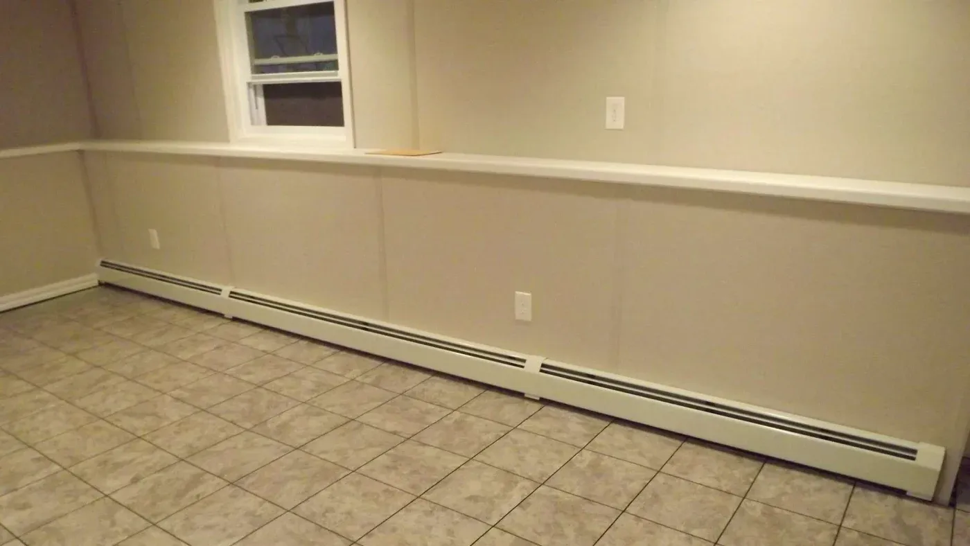 A tiled room with beige walls and a white baseboard heater running along the bottom. A small window and electrical outlets are visible. The space feels minimal and empty.