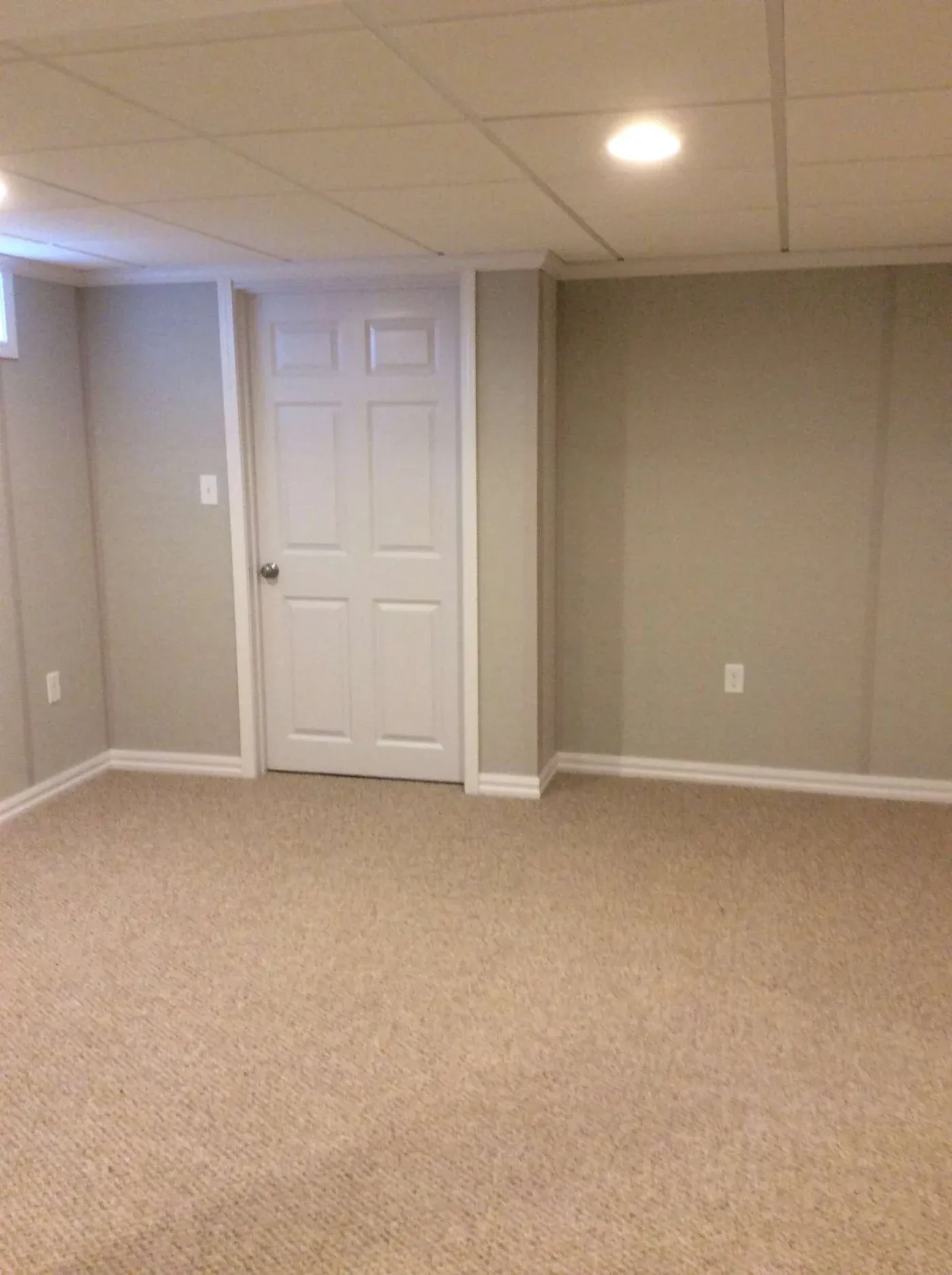 Finished basement room with beige carpet, white six-panel door, drop ceiling with recessed lighting, and neutral beige walls with electrical outlets