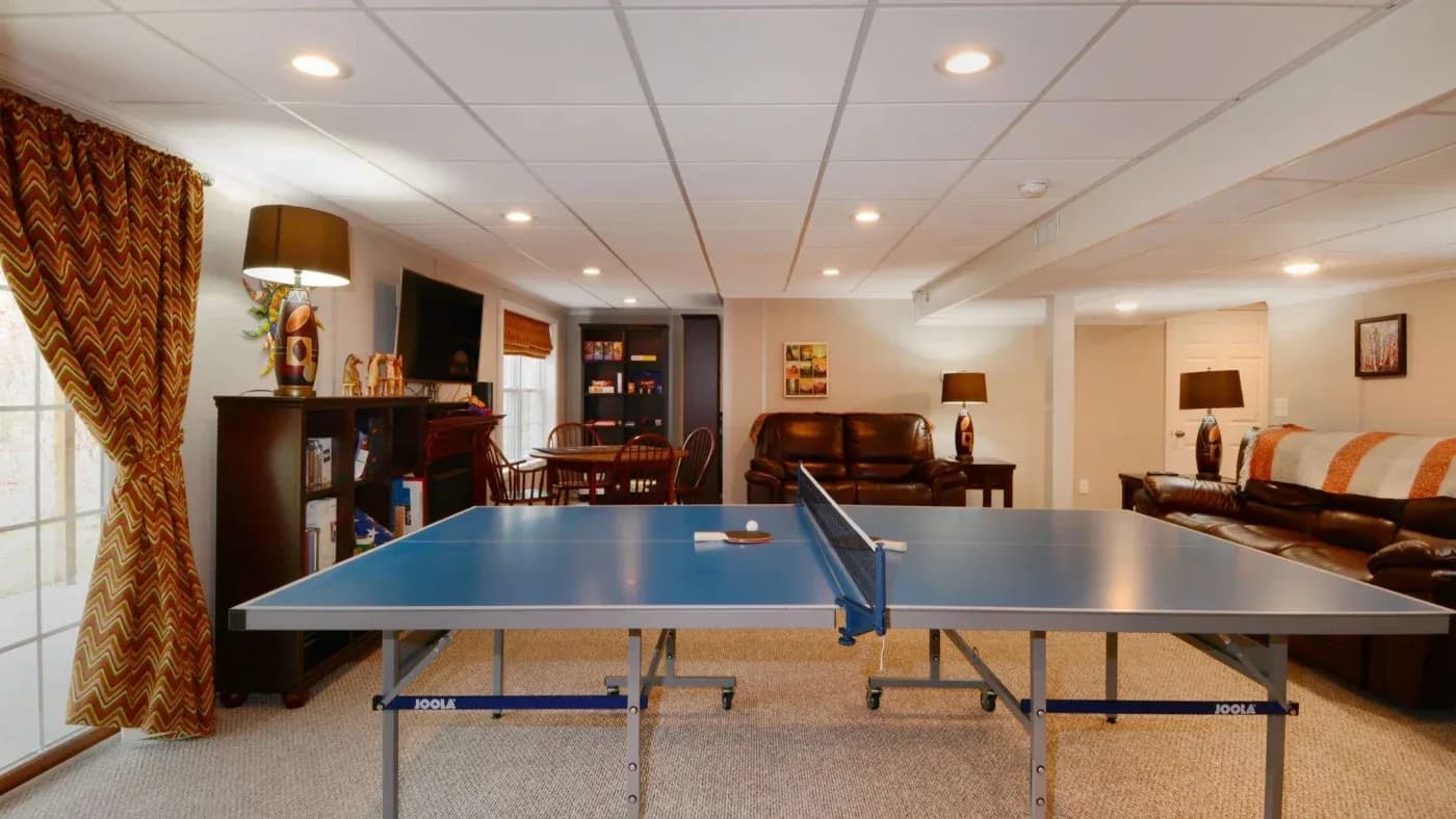 A cozy basement room features a blue ping pong table in the center, surrounded by brown leather couches and a wooden dining area, with warm lighting and patterned curtains creating an inviting atmosphere.