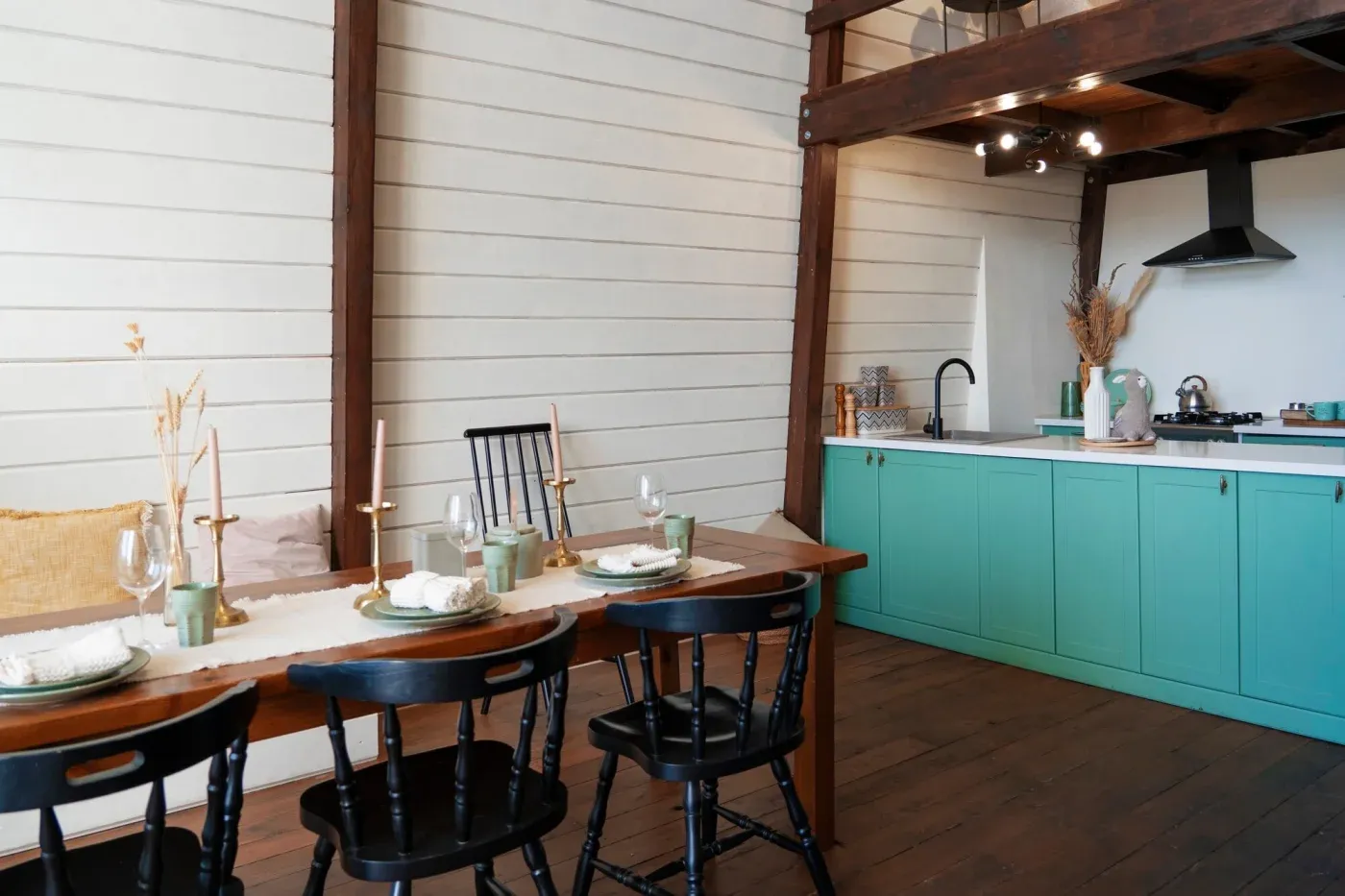 Cozy kitchen with wooden accents and teal cabinets. A rustic dining table with black chairs is set with plates and glasses, creating a warm, inviting atmosphere.