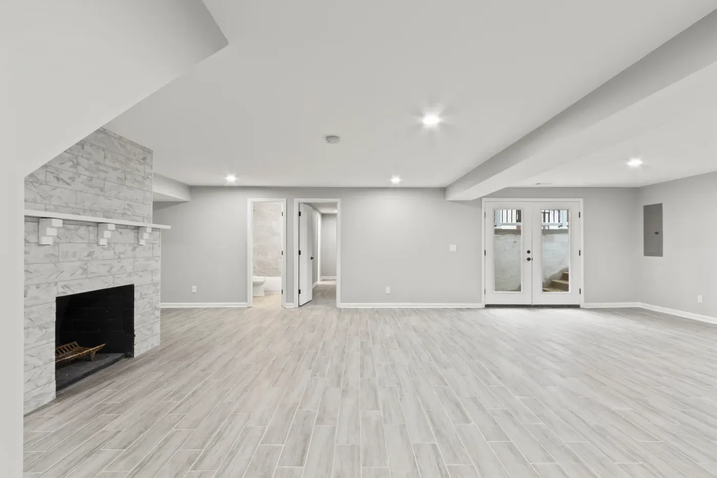Spacious basement with light gray wood flooring, a white brick fireplace, recessed lighting, and double doors leading outside. Minimalist and bright.