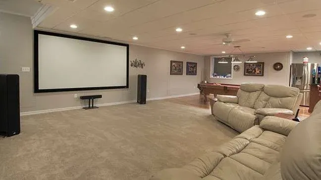 A cozy home theater room with a large projector screen on the wall, surround sound speakers, beige recliner sofas, and a pool table in the background.