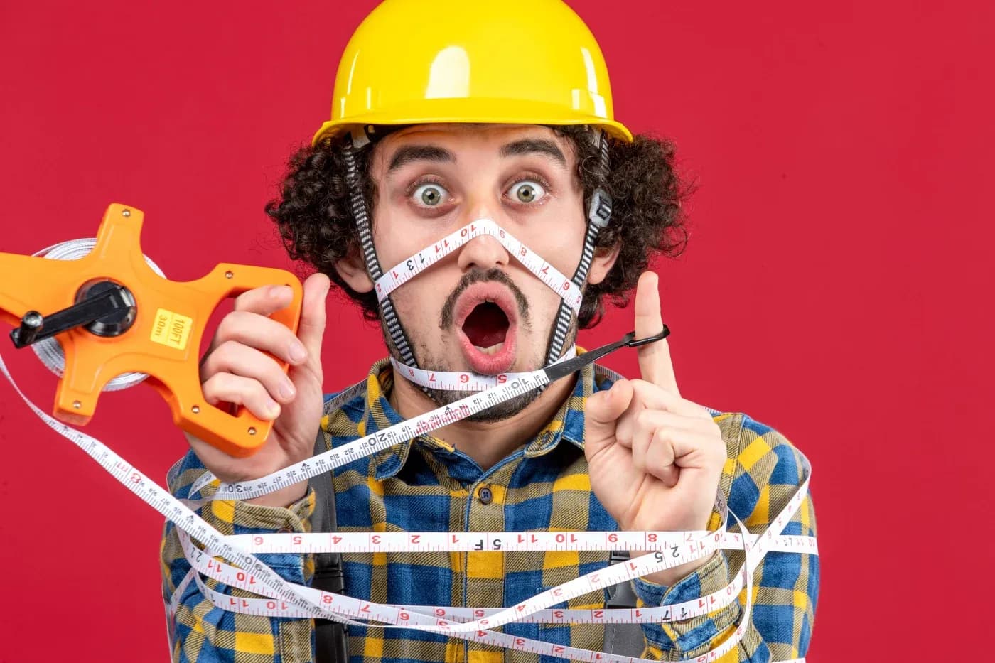A surprised man in a yellow hard hat, wrapped in measuring tape, holds an orange tape measure. He wears a plaid shirt against a red background.