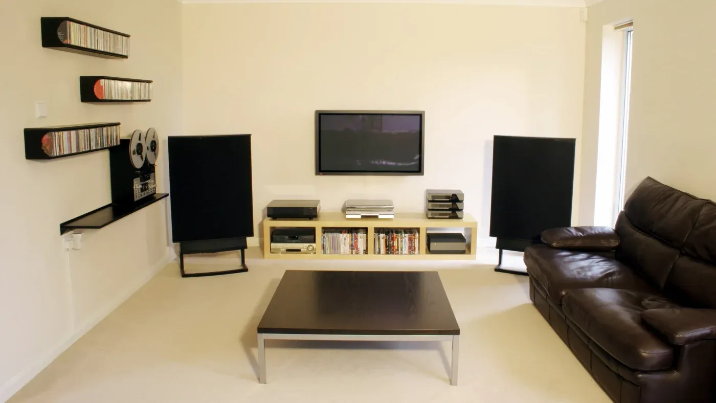 A modern living room with a cozy feel. It features a brown leather couch, flat-screen TV, speakers, and shelves with CDs. A dark wood coffee table sits in the center.