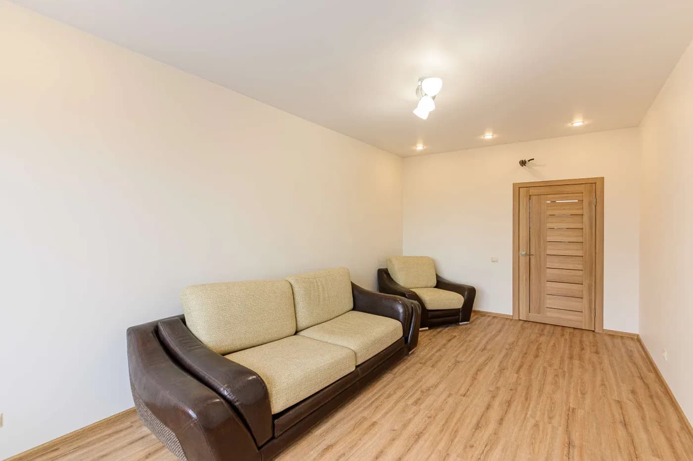 Minimalist room with light wood flooring, beige walls, and two brown sofas with cream cushions. A wooden door and ceiling lights add warmth.
