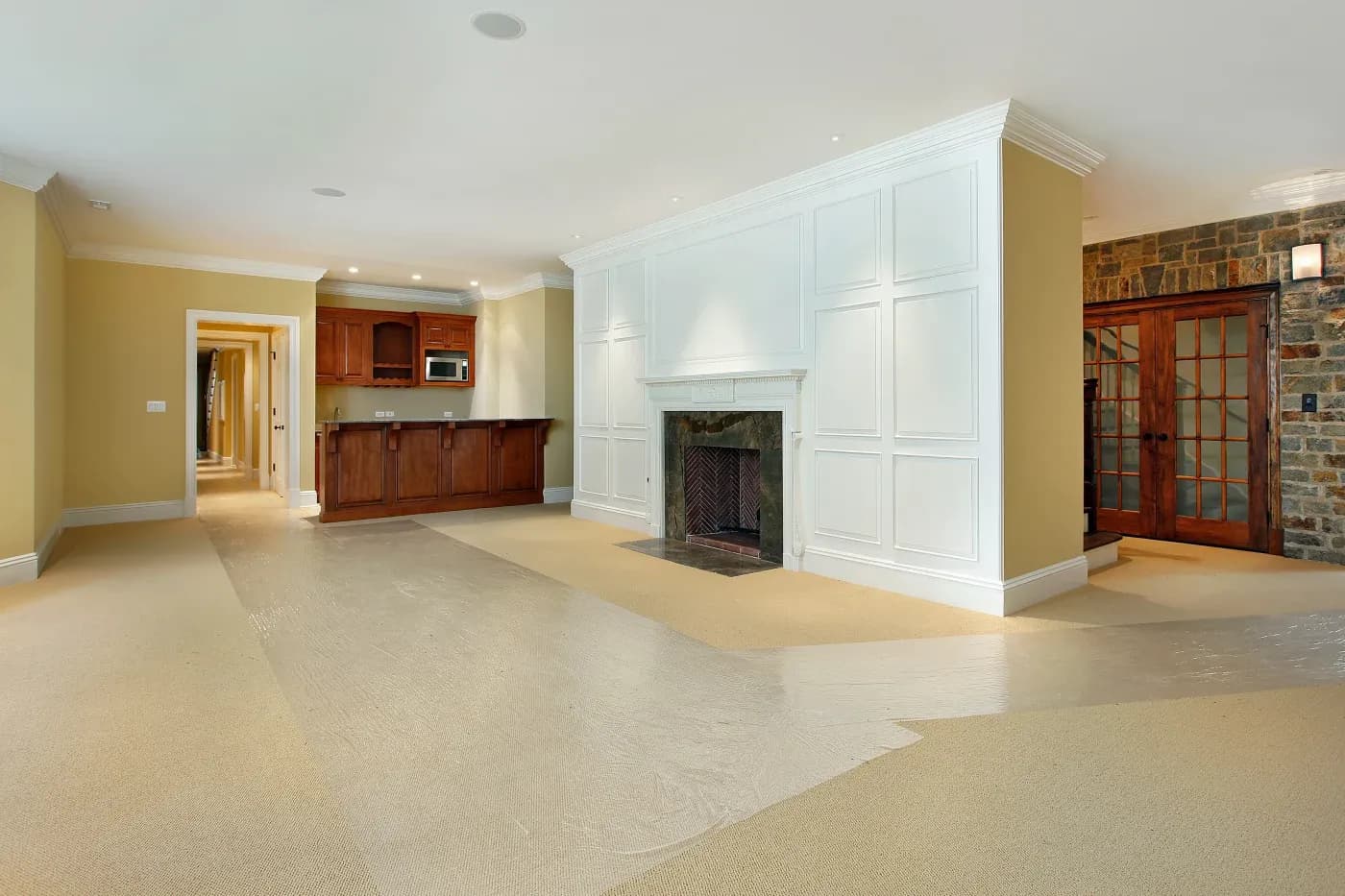 Spacious living room with beige carpet, white paneled fireplace, and built-in wooden mini bar. Stone accent wall with French doors adds a warm touch.