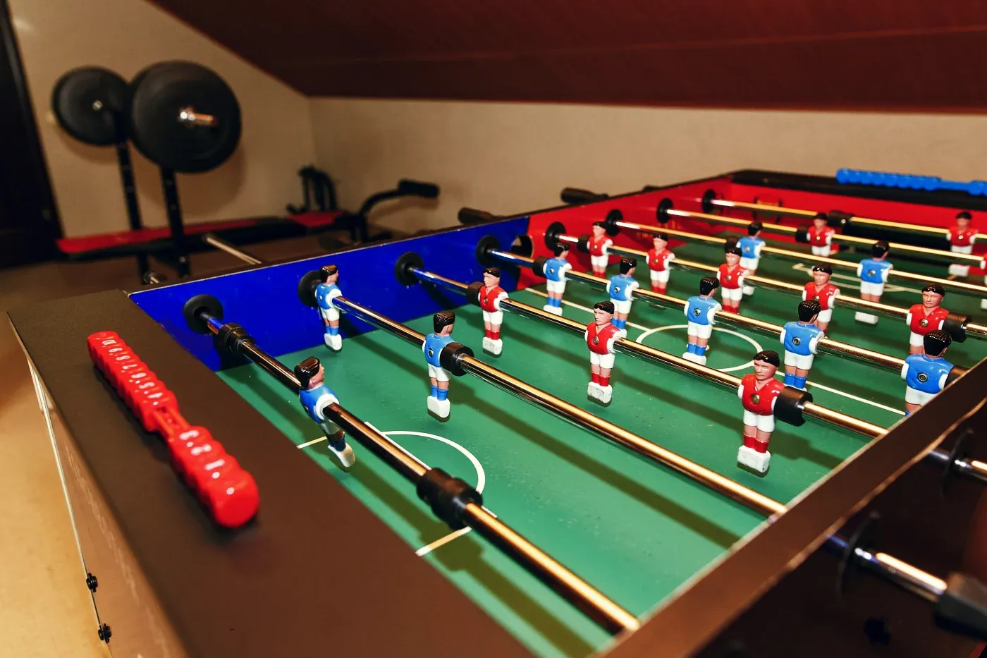 Foosball table game in a room with gym equipment, including weights. Red and blue player figures on a green field create an energetic, playful scene.