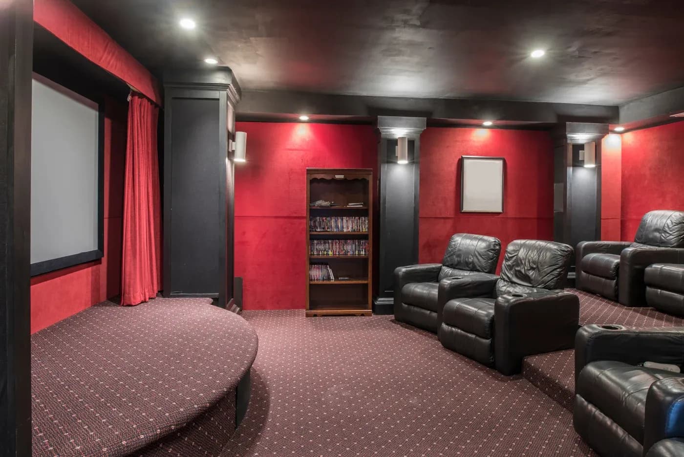 Home theater with black leather recliners, red walls, and dim lighting. A large screen is on the left, and a central shelf holds DVDs. Cozy ambiance.