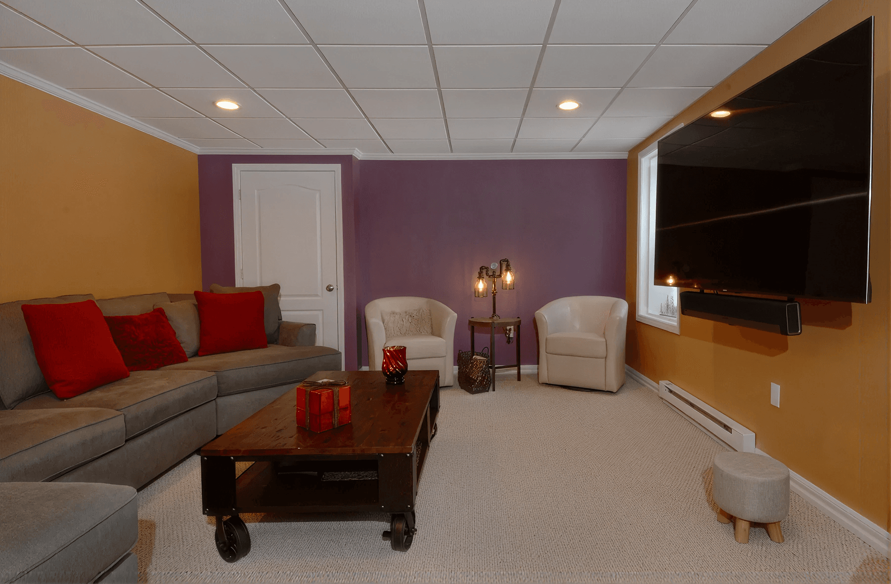 Cozy living room with a grey sectional sofa adorned with red cushions, a wooden coffee table, purple accent wall, and a large wall-mounted TV. Warm, inviting ambiance.