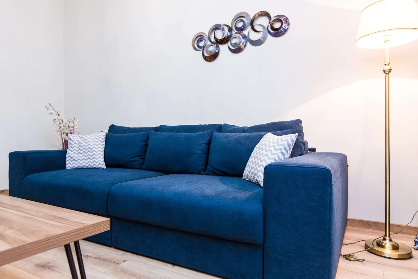 A cozy living room features a deep blue sofa with two chevron-patterned pillows. Above, metallic wall art; nearby, a tall lamp emits warm light.