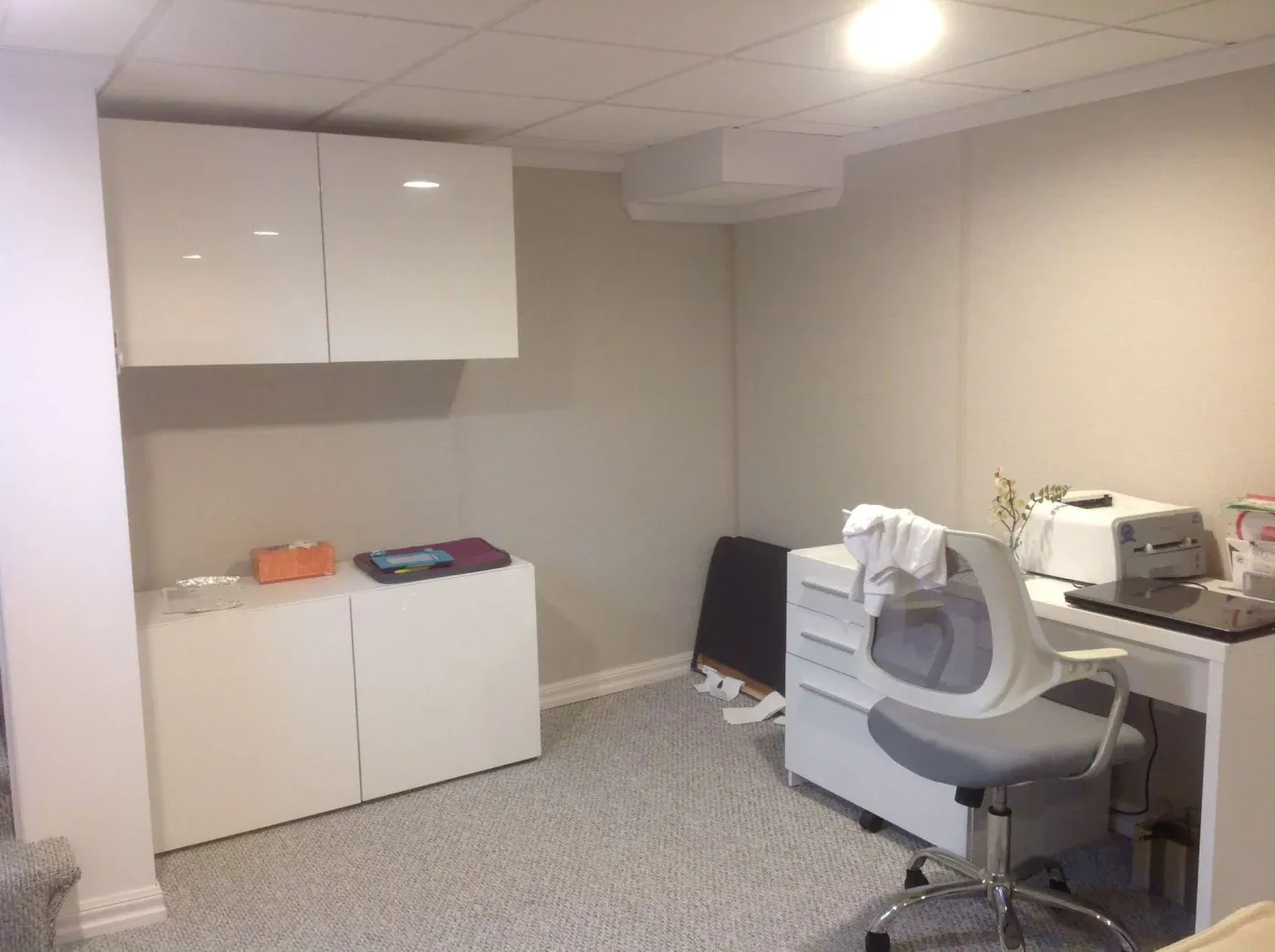Minimalist home office with white cabinets and desk, ergonomic chair, sewing machine, and soft lighting. The space is tidy and organized.