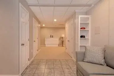 A cozy, softly lit room with beige walls and tiled floor. A gray sofa with a patterned cushion is on the right, near a white shelf with neatly arranged hats.
